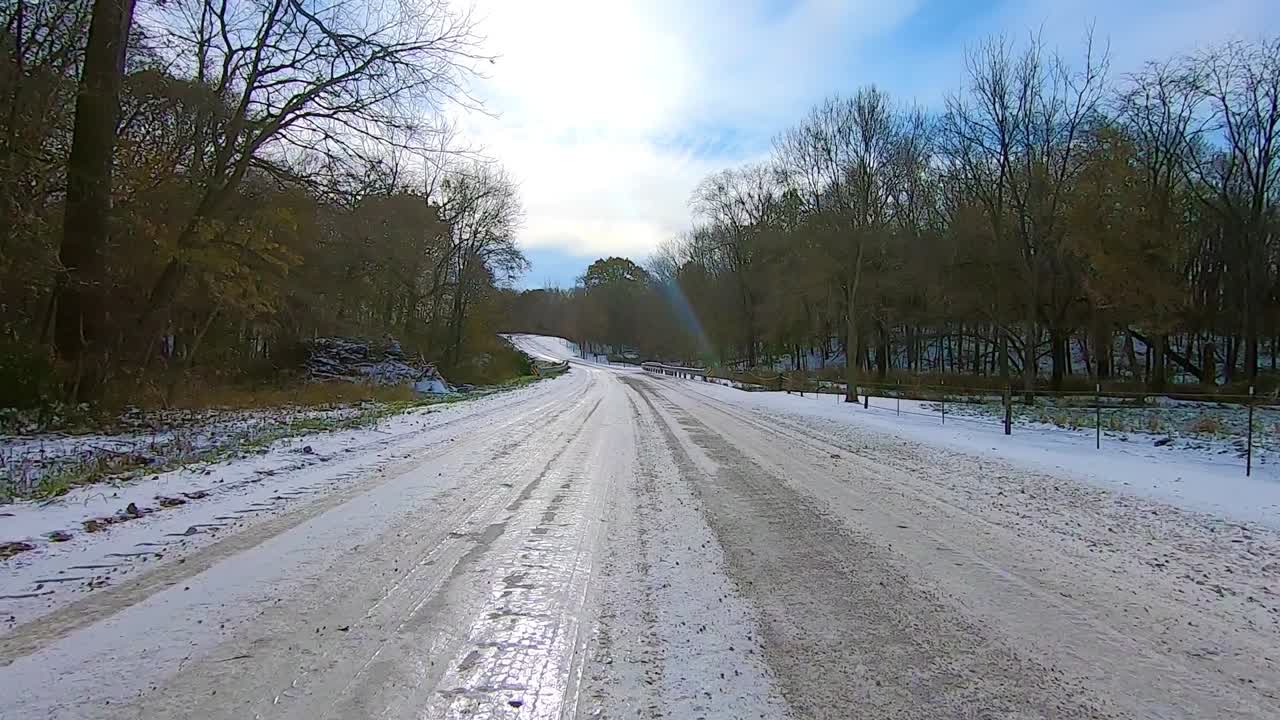 POV through the rear window while driving on a snowy and icy gravel road past timber and farm houses on a sunny winter afternoon; backwards Point of View