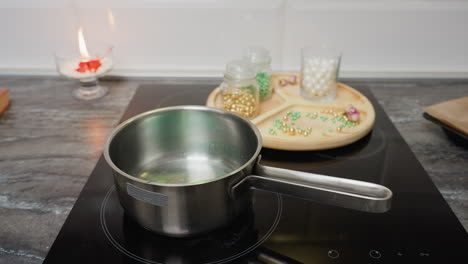 close-up of pot with green liquid on stovetop, beads of different colors on a wooden tray, and a burning red candle in the background