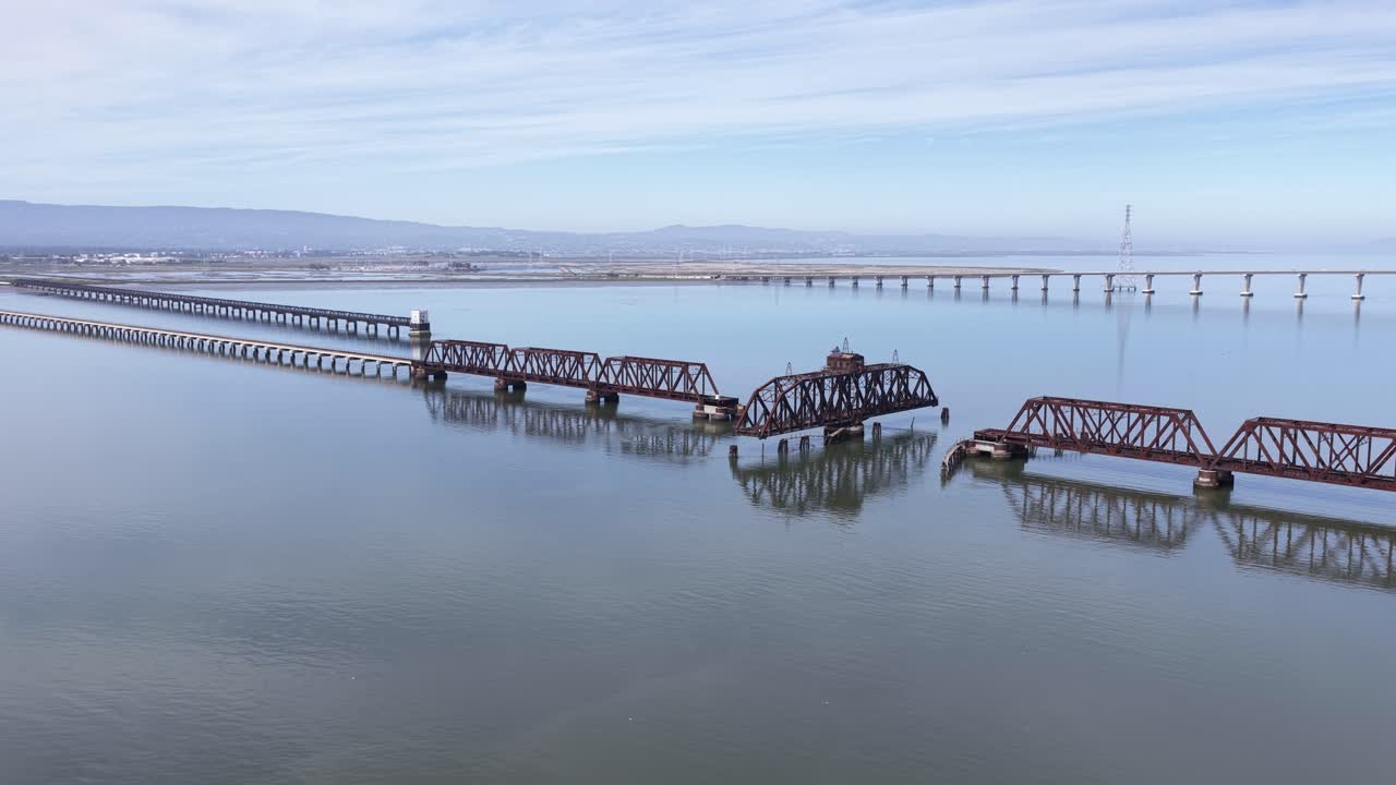 Vista da drone rotante sopra il ponte ferroviario di Dumbarton, abbandonato e arrugginito, un ponte girevole storico che attraversa la Baia di San Francisco, con l'acqua calma che riflette la sua struttura in decomposizione.