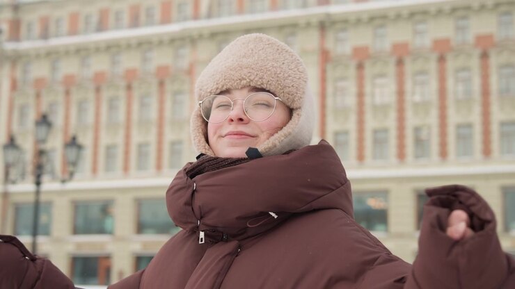 Primo piano di una donna che indossa un cappello di lana beige e un cappotto imbottito bordeaux con cappuccio foderato di pelliccia, che guarda in alto con un sorriso dolce mentre la neve cade in una piazza cittadina, un ritratto invernale calmo.