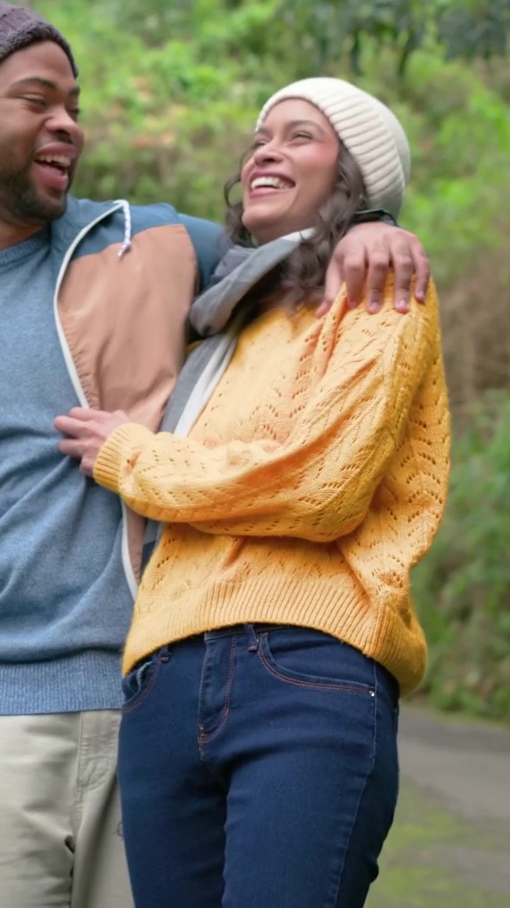 Video vertical: La cámara encuadra a una pareja caminando por un sendero boscoso, compartiendo risas, con gorros y bufanda