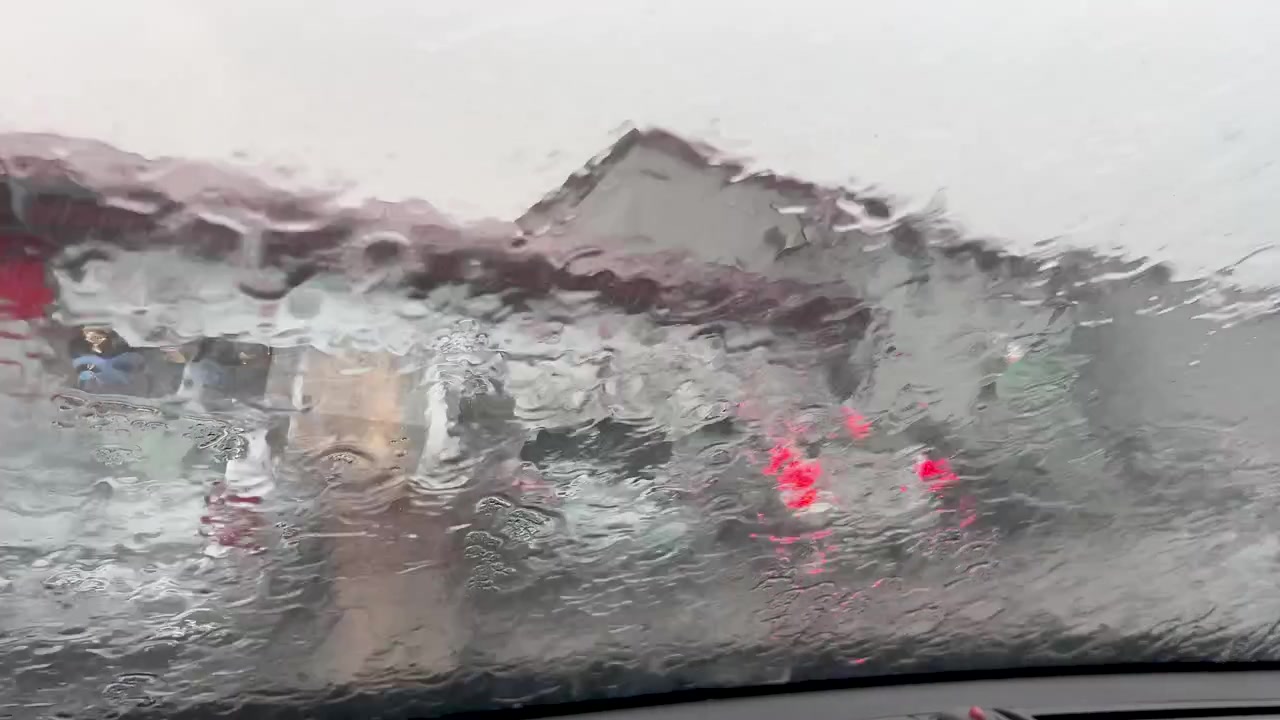 Close up of water droplets and heavy rain flowing down a vehicle windshield. Cinematic view of a stormy day from inside a car with blurred city background.