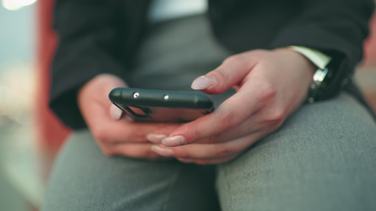 Close up of young lady with artificial nails holding and operating phone in both hands, wearing black jacket and wristwatch, with soft bokeh city lights in background