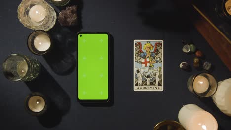 Overhead-Shot-Of-Person-Giving-Tarot-Card-Reading-With-Green-Screen-Mobile-Phone-Next-To-Judgement-Card-On-Table