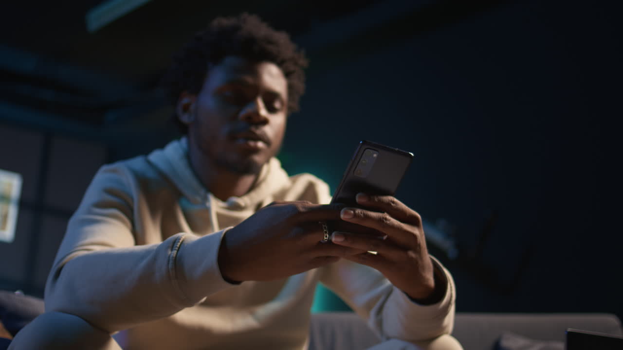 Relaxed black freelancer chatting with friends on his smartphone via texts, sitting on the couch. Young adult showcasing laughter and leisure, staying entertained while working remotely. Camera B.