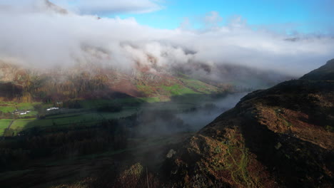 Flying-over-mountainside-towards-green-shadowed-valley-and-cloud-shrouded-mountains-lit-by-early-morning-autumn-sunshine