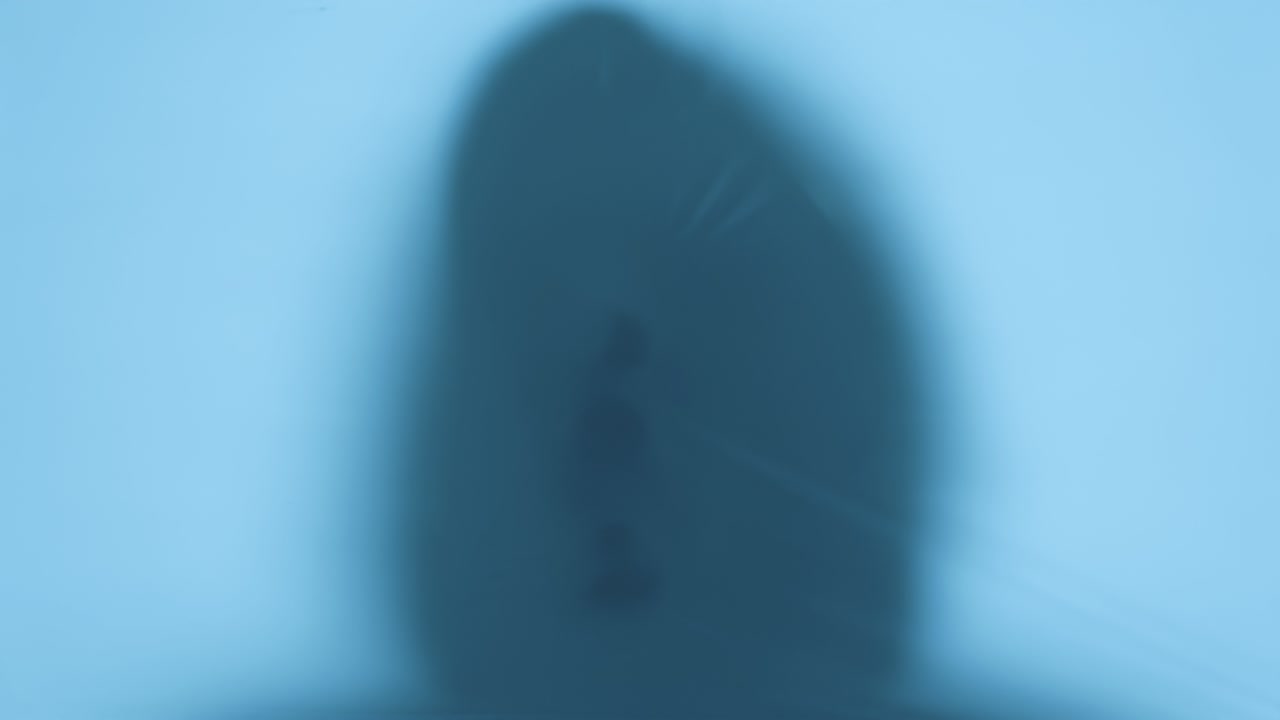 Close-up silhouette of a hooded human's head breaking through translucent cellophane. Ideas about the other world, victims of violence, paronormality, ghosts. 4k video loop with speed ramp effect.