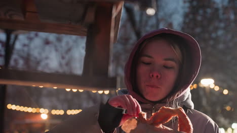 young woman standing outdoors at dusk in snowy park, gently placing food into bird house while holding food in hand, surrounded by snow-covered ground, soft street lights in background