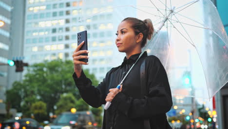 Woman-standing-with-a-clear-umbrella,-using-her-smartphone-in-the-rain,-with-a-cityscape-background