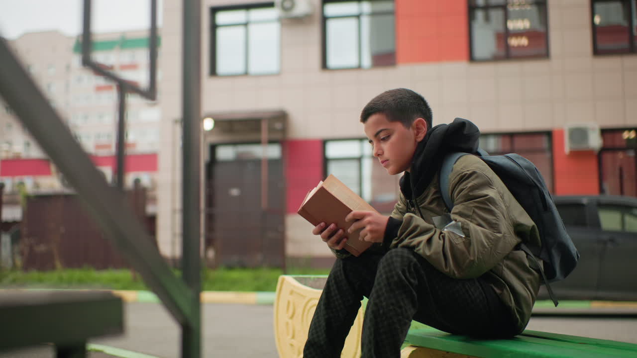 Vista lateral de un estudiante concentrado hojeando las páginas de una novela mientras está sentado al aire libre con un edificio borroso al fondo, mostrando concentración