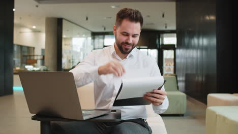 smiling young businessman using laptop and analyzing graph documents reports in modern office lobby