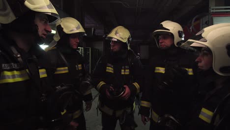 group of five firefighters wearing full equipment in smokey area. relaxed chatting after work well done. fire trucks in the background. name tag translation is fireman and model's last name.