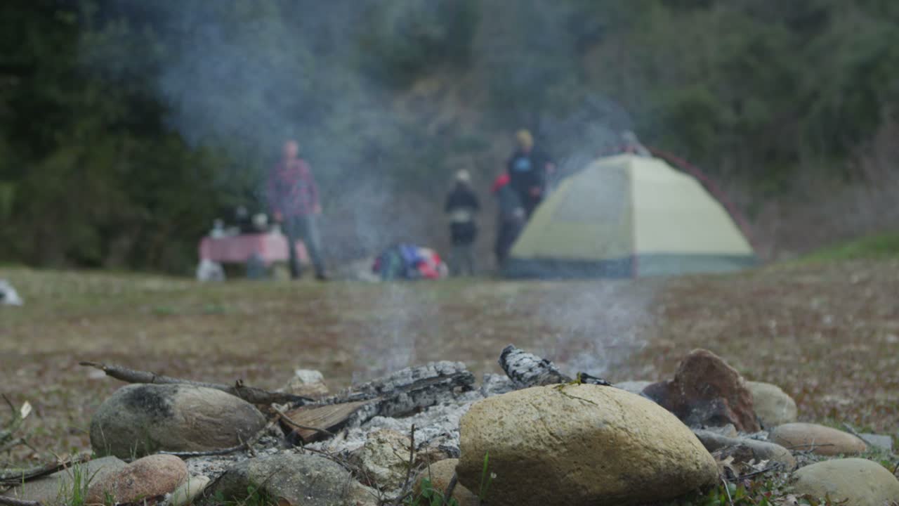 캠프 참가자들이 텐트 근처에서 배경으로 이동하는 동안 꺼진 모닥불 연기