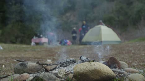 an extinguished campfire smolders while campers move in the background near a tent