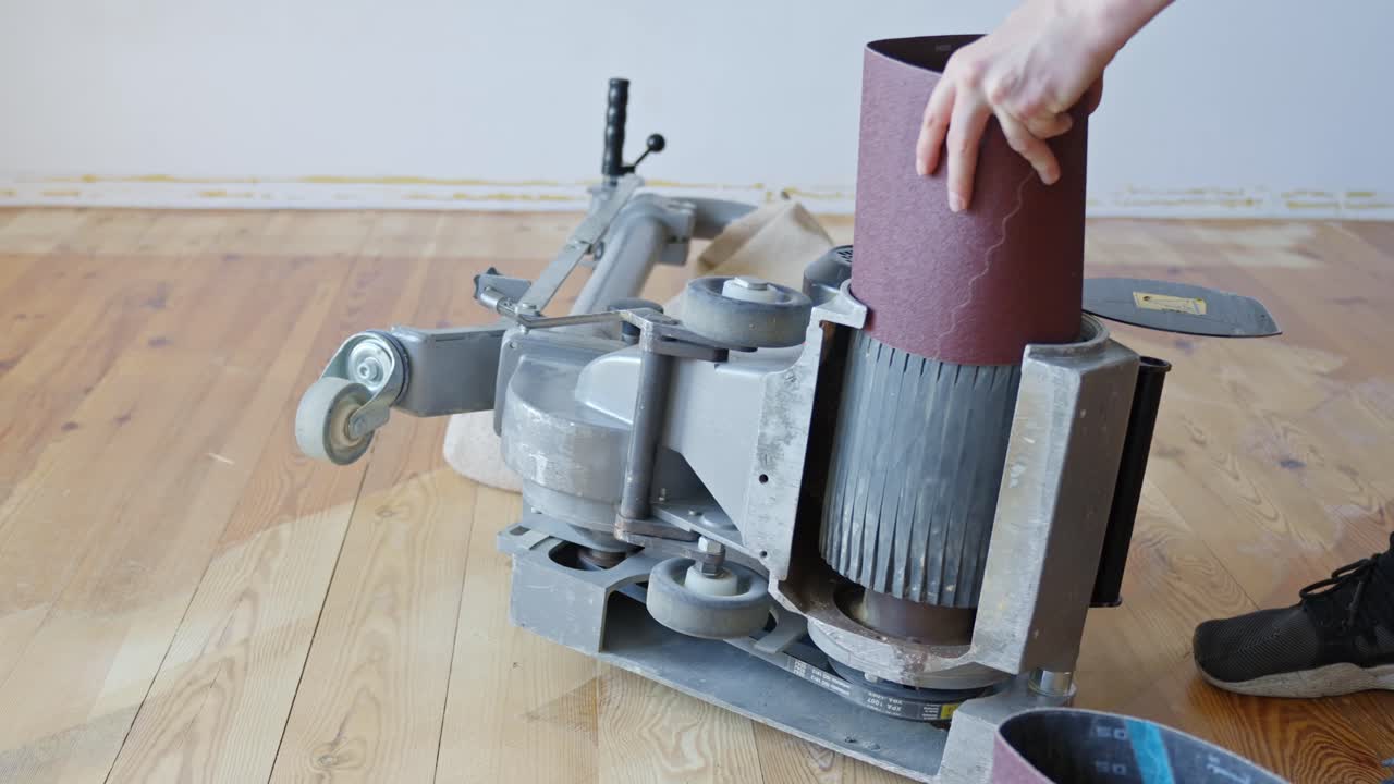 Person loads a fresh sandpaper roll onto a professional wood floor sanding machine positioned on pine floor, preparing the equipment for surface restoration, renovation work, static camera, real time