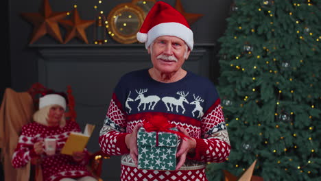 Elderly-grandfather-in-festive-sweater-presenting-Christmas-gift-box,-smiling,-looking-at-camera