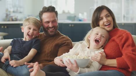 Familia-Sentada-En-El-Sofá-Con-Palomitas-De-Maíz-Riendo-Viendo-Comedia-En-La-Televisión-Juntos