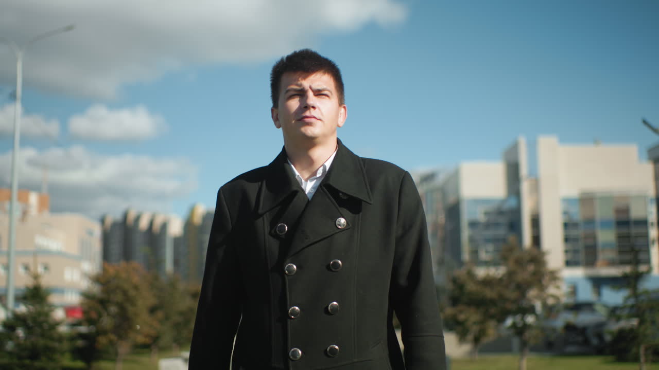 검은 코트를 입은 남자가 은 파란 하늘 아래 현대적인 사무실 건물, 움직이는 자동차, 그리고 활기찬 나무가 있는 바쁜 도시 도로를 따라 자신감 있게 고 있습니다.