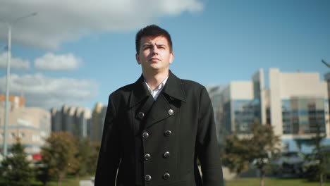 man in black coat walking confidently along busy urban road with modern office buildings, moving cars, and vibrant trees in background under clear blue sky
