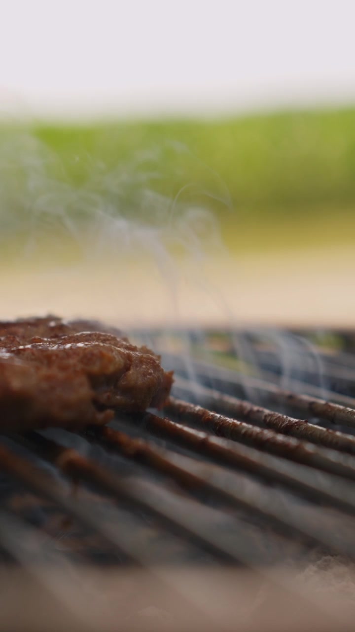 Video verticale ravvicinato di un succulento hamburger di manzo che sfrigola su una griglia calda all'aperto, con fumo aromatico che sale per gustose storie social e utilizzo su dispositivi mobili