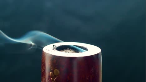 static shot of a antique vintage pipe being stationary and burning producing white blue smoke looking like a spirit moving away to the sides and above smoking tobacco wooden black background