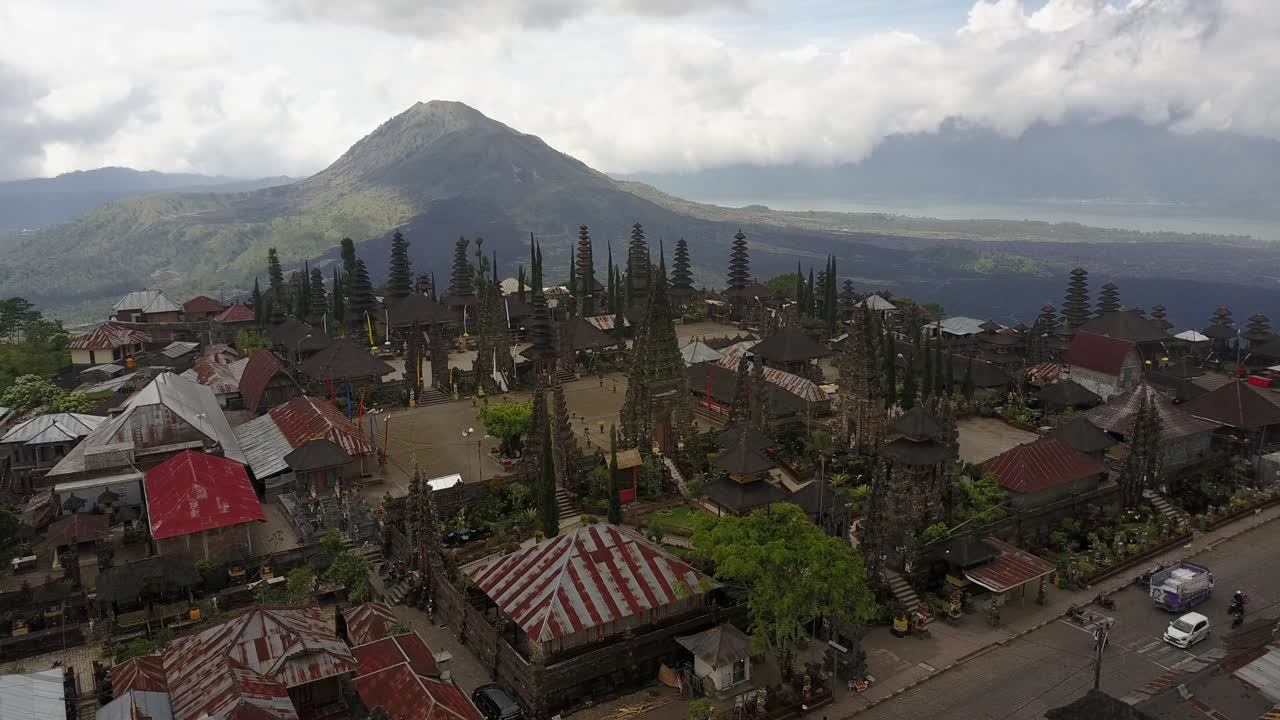 Klip drone udara terbang maju menuju Pura Besakih, candi induk Bali, dengan gunung berapi Gunung Agung menjulang di latar belakang, menunjukkan arsitektur tradisional dan kehidupan sehari-hari di Bali, Indonesia