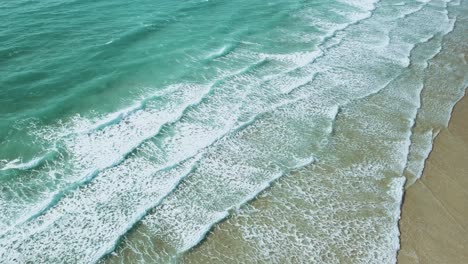 Wide-aerial-shot-of-crystal-clear-waves-crashing-onto-a-sandy-beach