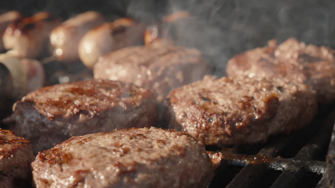 Hamburger di manzo in cottura e sfrigolanti su barbecue fumante con salsicce di maiale sullo sfondo