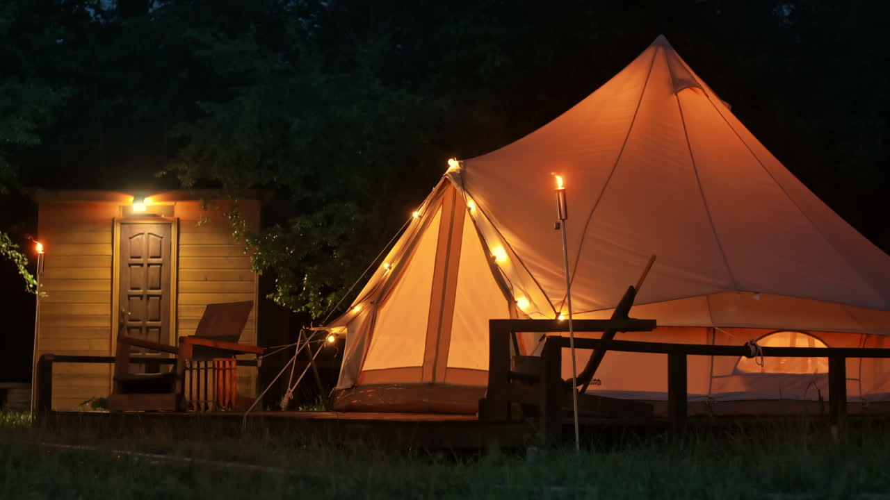 Telt med brændende fakler, lamper og træstole ved glamping, skov rundt om, nat