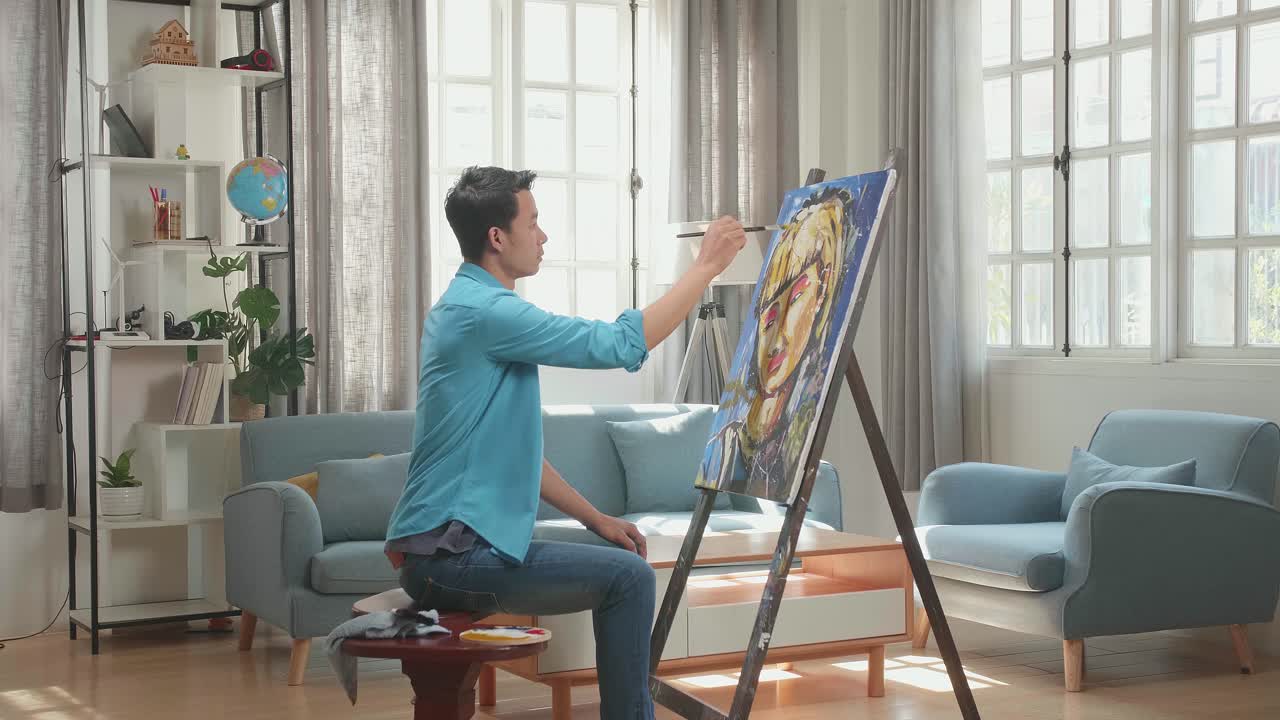 Asian Artist Man Sitting On Round Wooden Chair Without Backrest Holding Paintbrush Mixed Colour And Painting A Girl'S Hair On The Canvas
