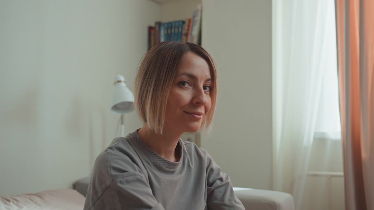 Donna caucasica amante della musica seduta sul divano, luce diurna soffusa attraverso la finestra, libreria e lampada visibili dietro, maglietta grigia casual e taglio bob corto, sorriso pensieroso e postura rilassata, atmosfera intima.