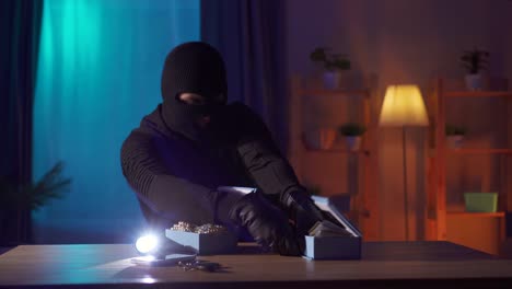 un ladrón con una balaclava negra encuentra joyas y dinero en una mesa de la casa.