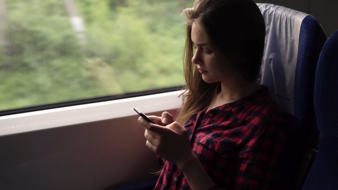 chica joven seria y relajada sentada en el tren cerca de la ventana. viajando en un tren moderno. chica de pelo largo está desplazándose o escribiendo en su teléfono móvil. camisa roja a cuadros. vista lateral