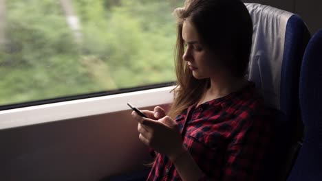 Una-Joven-Seria-Y-Relajada-Sentada-En-El-Tren-Cerca-De-La-Ventana.-Viajando-En-Tren-Moderno.-Una-Chica-De-Pelo-Largo-Se-Desplaza-O-Escribe-En-Su-Teléfono-Móvil.-Camisa-Roja-A-Cuadros.-Vista-Lateral