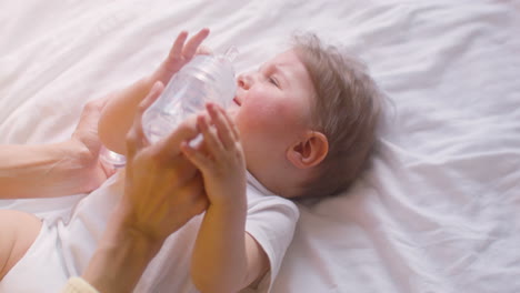 top view of a crying baby lying on the bed in bedroom while his mother is trying to give him water with the feeding bottle