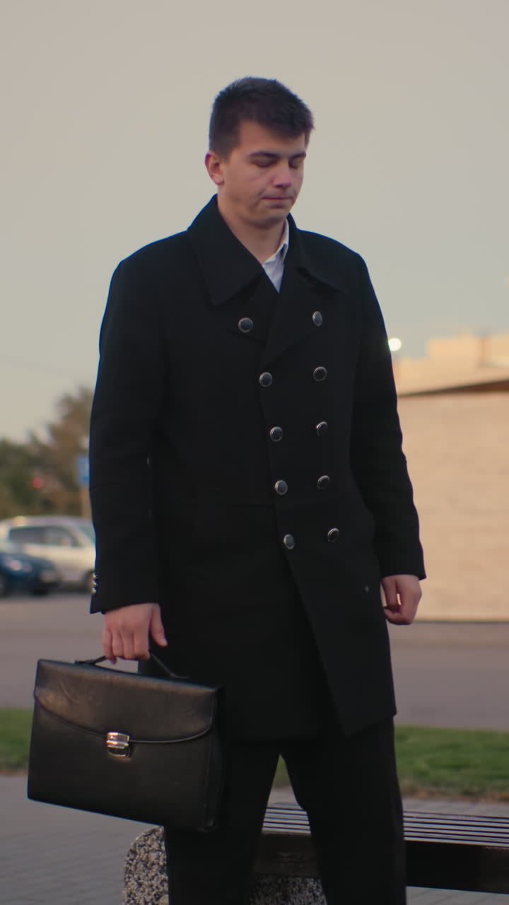 hombre de traje negro camina hasta el banco, abre el maletín en un entorno al aire libre con edificios modernos y coches estacionados, coche en movimiento en el fondo al amanecer, luz de la calle brillando temprano en la mañana