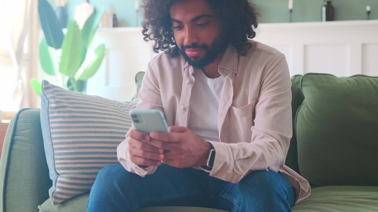 Giovane uomo arabo millennial usa il telefono cellulare seduto sul divano in casa