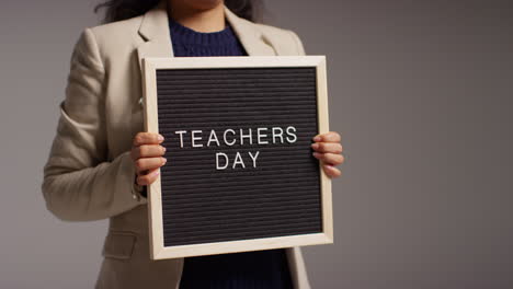 Close-Up-Studio-Shot-Of-Female-Teacher-Standing-Against-Grey-Background-Holding-Notice-Board-Reading-Teachers-Day-3