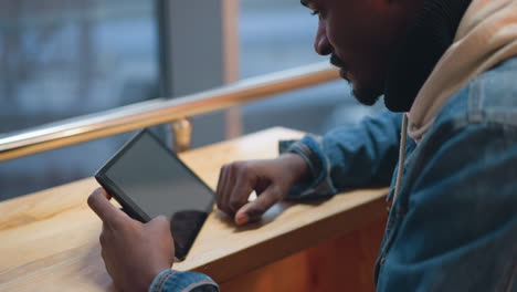 close up of young black male holding tablet at wooden counter by large window overlooking cityscape dusk inside modern cafe interior wearing denim jacket and hoodie casual relaxed vibe scene