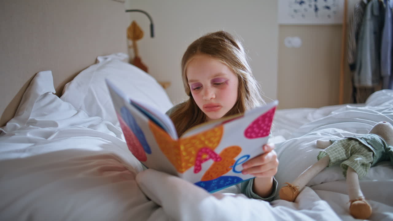 Focused kid reading book resting in cozy bedroom. Makeup small girl lying bed