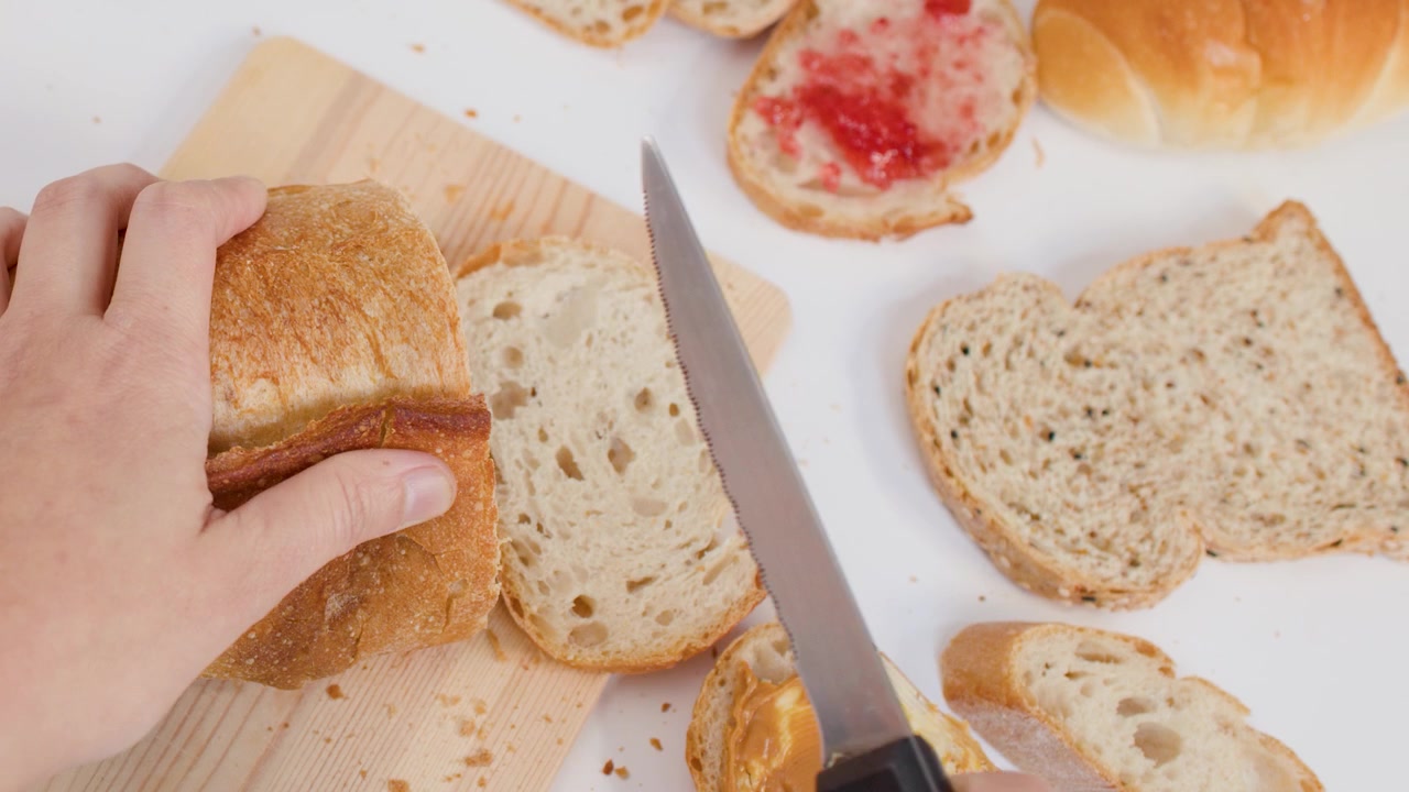 Ripresa dall'alto di mani che tagliano pane croccante con un coltello su un tagliere accanto a pane tostato con marmellata.