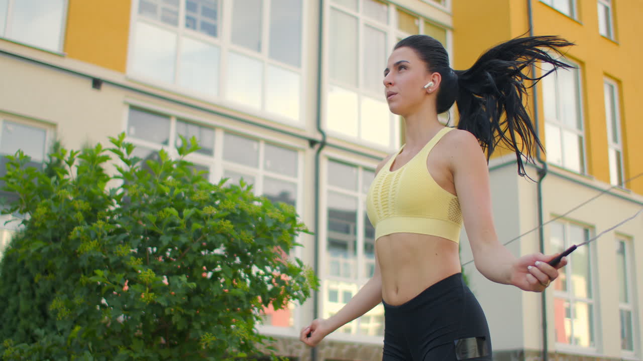 젊은 아름다운 소녀는 스포츠 옷을 입고 슬로우 모션으로 공원에서 점프 로프에 점프합니다. 집의 배경에 스포츠를 재생