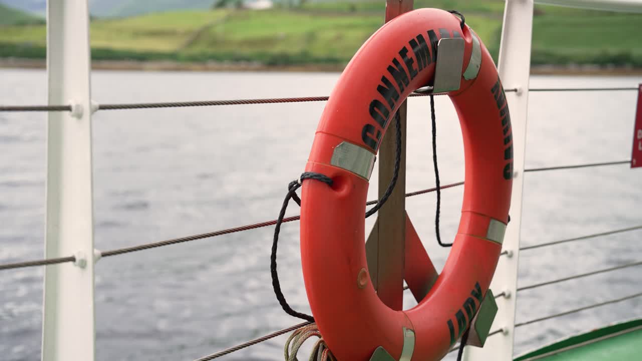 Close-up of an orange lifebuoy on the 'Connemara Lady' tour boat. The vessel moves through the water with the scenic green hills of Connemara, Ireland, in the background