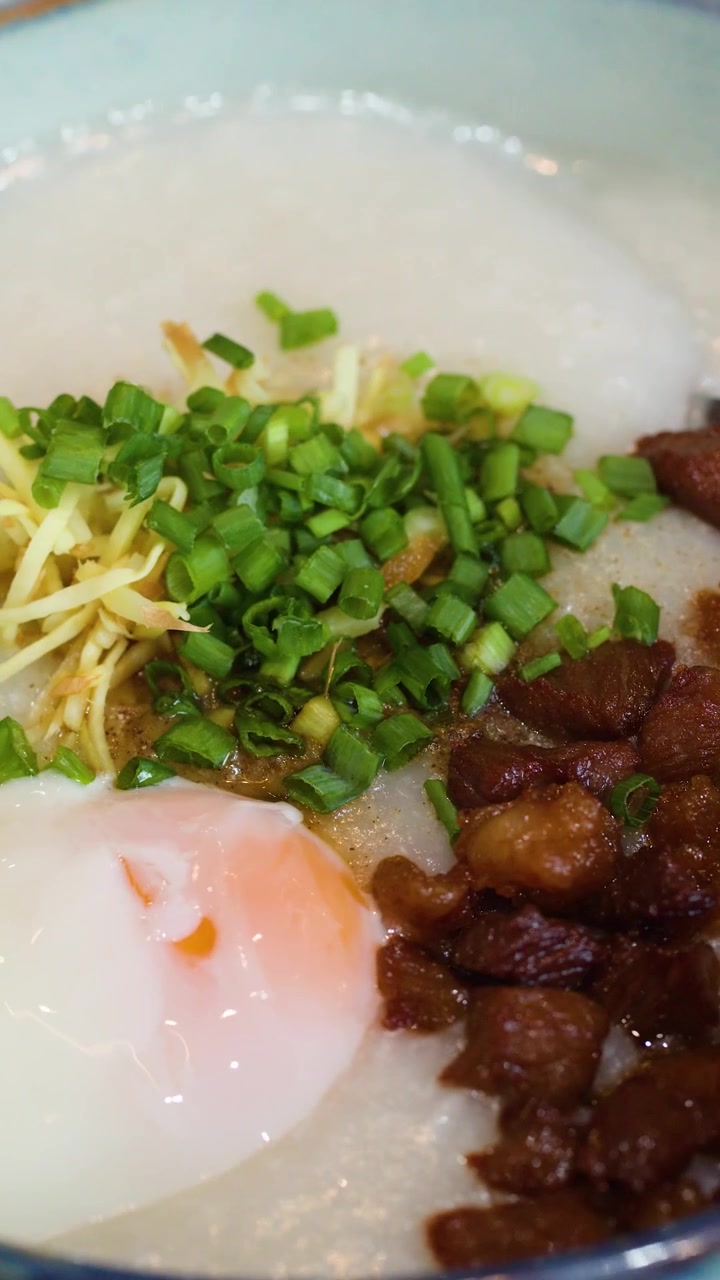 Close-up bubur nasi tradisional Asia dengan topping telur, daging babi, dan daun bawang.