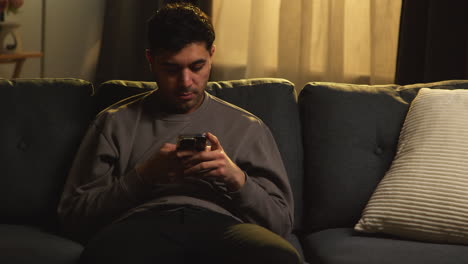 young man spending evening at home sitting on sofa with mobile phone scrolling through internet or social media 1