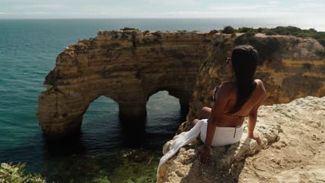 a woman relaxes on a cliffside overlooking natural sea arches and turquoise waters on a sunny day in albufeira, portugal