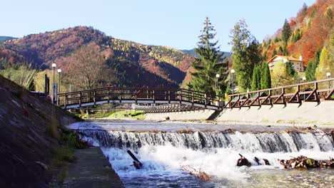 small waterfall on river with bridge and mountain in background in piatra craiului mountains, brasov, romania during autumn