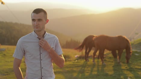 Young-man-is-standing-in-the-field-near-mountains.-Behind-him-grazing-horses