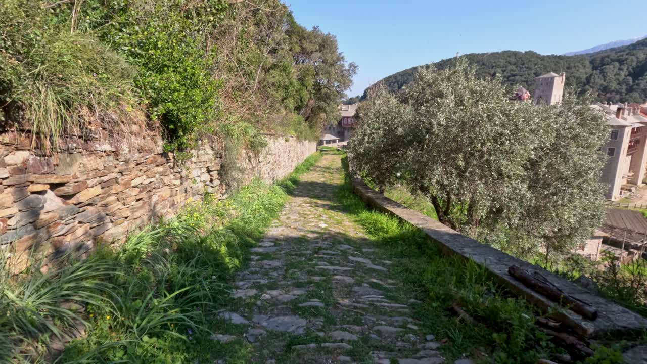 Una persona camina por un sendero de piedra rodeado de vegetación, árboles y muros. El sol brilla y un pueblo es visible a lo lejos.