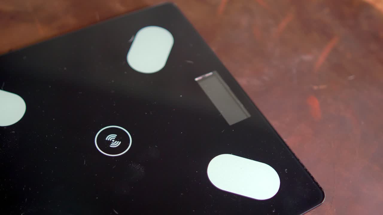 A person's feet in black socks stand on a black digital scale with a blue light, placed on a scratched wooden floor indoors